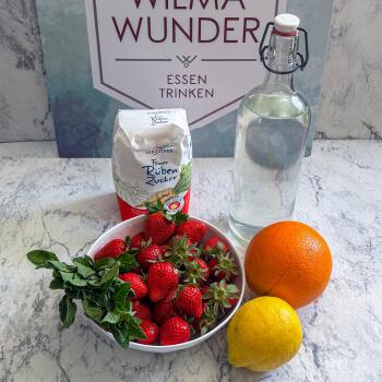 Tisch mit einer Schüssel voll Erdbeeren und einem Bund Minze, daneben eine Orange, eine Zitrone, Rohrzucker und eine Flasche Wasser.