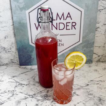 Glas auf einem Tisch stehend, Limonade mit Erdbeere-Minz-Syrup und einer Zitrone am Rand. Daneben eine Flasche mit dem Sirup auf Marmor Tisch und Hintergrund.